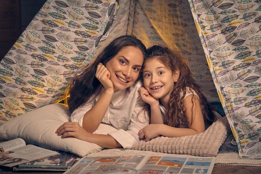 Mother and daughter are sitting in a teepee tent with some pillows and reading stories. They are smiling and looking at the camera. Happy family.