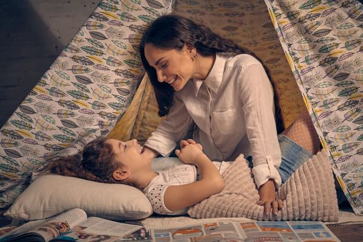 Mother and her daughter are in a teepee tent with some pillows. They are smiling and looking at each other. Happy family.