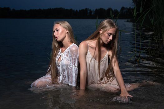 two attractive young twin sisters with long blond hair posing in light dresses in water of lake at summer night. stylish fashion photoshoot with flashlight. pretty models outdoors evening photosession