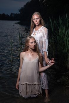 two attractive young twin sisters with long blond hair posing in light dresses in water of lake at summer night. stylish fashion photoshoot with flashlight. pretty models outdoors evening photosession