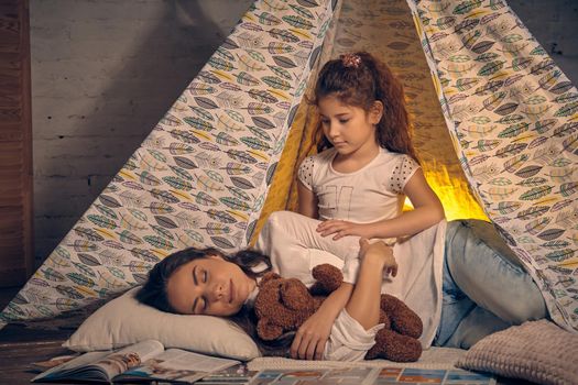 Young mother and her pretty daughter are in a teepee tent with some pillows. Mom is sleeping with a toy bear. Daughter is looking at her. Happy family.