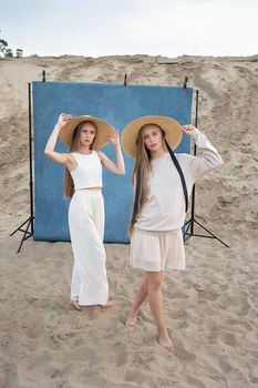 beauty portrait outdoors on sand in front of blue background, young pretty twins with long blond hair posing in sand quarry in elegant white, beige clothes. identical caucasian sisters pose in hats