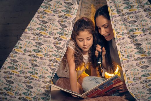 Mother and her beautiful daughter are sitting in a teepee tent with some pillows, reading stories with the flashlight. Happy family.