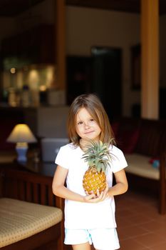 Little caucasian female kid standing in living room and keeping pineapple. Concept of fruit, vegeterian life and childhood.