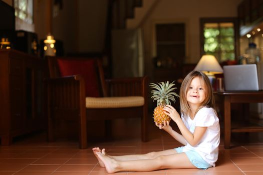 Little nice female kid sitting on floor in living room and playing with pineapple, laptop in background. Concept of health life, fruit and childhood.