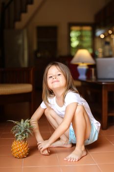 Little smiling barefoot girl playing with pineapple and sitting on floor. Concept of health vegeterian lifestyle and children.