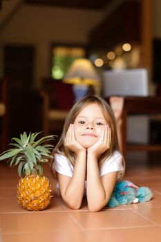 Portrait of little girl lying on floor and playing with pineapple and toy, laptop in background. Concept of health vegeterian life and childhood.
