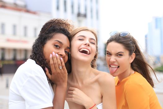 Three young smiling hipster women in summer clothes posing on street.Female showing positive face emotions