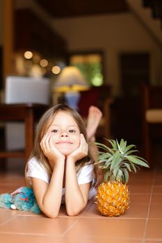 Portrait of little cute girl lying on floor and playing with pineapple and toy, laptop in background. Concept of health vegeterian life and childhood.