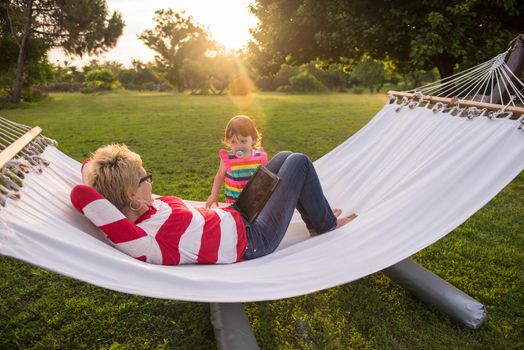 Happy mother and her little daughter enjoying free time using tablet computer while relaxing in a hammock during sunny day on holiday home garden