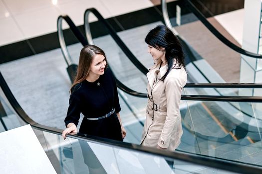 two business women talking standing on an escalator in a business center