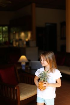 Little female kid standing in living room and keeping pineapple. Concept of fruit, vegeterian life and childhood.