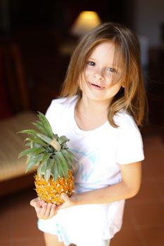 Portrait of little female kid standing in living room and keeping pineapple. Concept of fruit, vegeterian life and childhood.