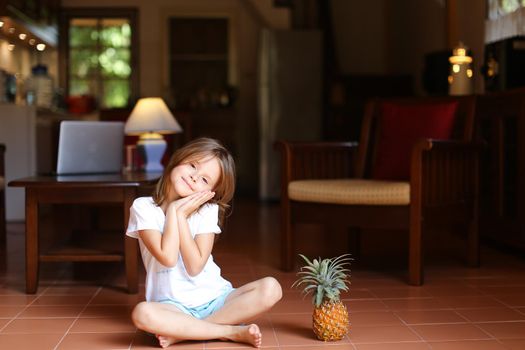 Little female kid sitting on floor in living room and keeping pineapple, laptop in background. Concept of health life, fruit and childhood.