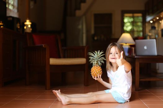 Little smiling female kid sitting on floor in living room with pineapple and showing thumbs up, laptop in background. Concept of health life, fruit and childhood.