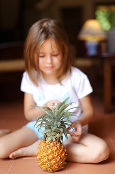 Closeup pineapple with little girl sitting barefoot on floor in background. Concept of health life, fruit and kids.