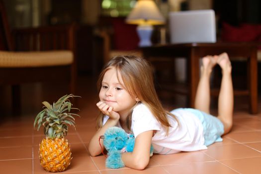 Little barefoot girl lying on floor and playing with pineapple and toy, laptop in background. Concept of health vegeterian life and childhood.