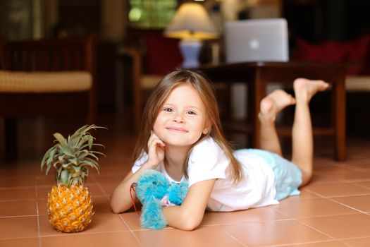 Little european girl lying on floor and playing with pineapple and toy, laptop in background. Concept of health vegeterian life and childhood.