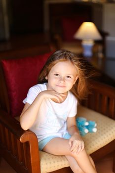 Little beautiful female kid sitting with toy in wooden chair. Concept of kid model and wood in interior.