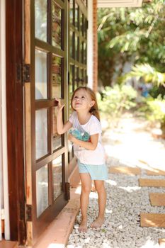 Little american female kid standing near door with toy outside, palms in background. Concept of resting with kids in Phuket, exotic hotel.