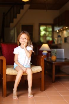 Female happy kid sitting in wooden chair in living room. Concept of child model and cozy interior.