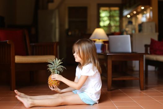 Little female blonde kid sitting on floor in living room and keeping pineapple, laptop in background. Concept of health life, fruit and childhood.