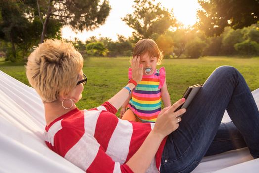 Happy mother and her little daughter enjoying free time using tablet computer while relaxing in a hammock during sunny day on holiday home garden