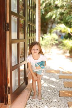 Little european female kid standing near door with toy outside, palms in background. Concept of resting with kids in Phuket, exotic hotel.