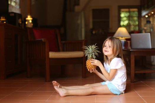 Little cute female kid sitting on floor in living room and playing with pineapple, laptop in background. Concept of health life, fruit and childhood.