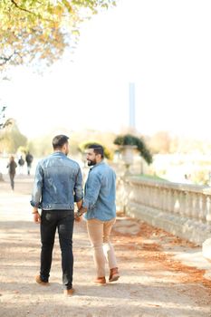 Back view of two caucasian gays walking in park and holding hands, wearing jeans shirt. Concept of same sex couple and relationship.