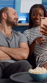 Modern interracial couple talking on video call conference at home. Young multi ethnic partners using internet for remote communication, sitting together on couch in living room.