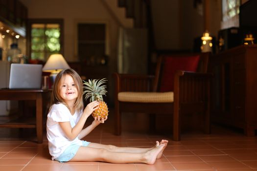 Little smiling female kid sitting on floor in living room and playing with pineapple, laptop in background. Concept of health life, fruit and childhood.