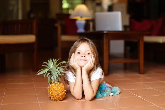 Little caucasian girl lying on floor and playing with pineapple and toy, laptop in background. Concept of health vegeterian life and childhood.