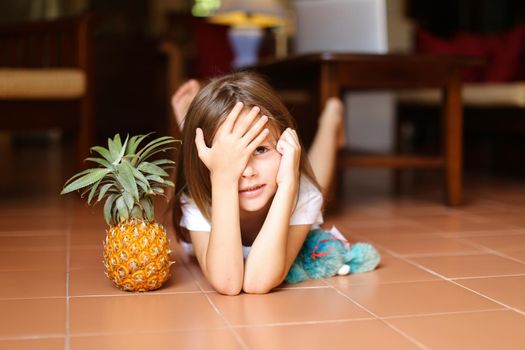 Portrait of little nice girl lying on floor and playing with pineapple and toy, laptop in background. Concept of health vegeterian life and childhood.
