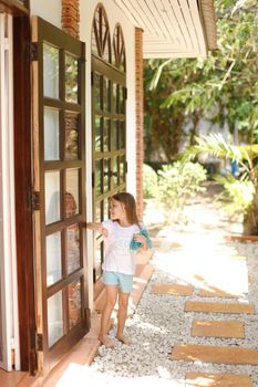 Little female kid standing near door with toy outside, palms in background. Concept of resting with kids in Phuket, exotic hotel.