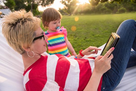 Happy mother and her little daughter enjoying free time using tablet computer while relaxing in a hammock during sunny day on holiday home garden