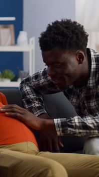 Pregnant interracial couple sitting on living room couch. African american husband touching baby bump and talking to child while caucasian mother relaxing on sofa and laughing