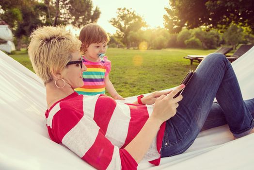Happy mother and her little daughter enjoying free time using tablet computer while relaxing in a hammock during sunny day on holiday home garden