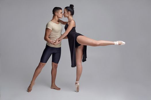 Pair of a graceful ballet dancers are posing over a gray studio background and looking at each other. Handsome man in black shorts with beige t-shirt and beautiful woman in a black dress and white pointe shoes are dancing together. Ballet and contemporary choreography concept. Art photo.