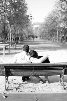 Black and white photo, back view two hugging gays sitting on bench in park. Concept of same sex couple and resting outside.