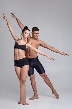 A pair of modern ballet dancers in black suits are posing over a gray studio background. Handsome man in black shorts and beautiful woman in a black swimwear are dancing together. Ballet and contemporary choreography concept. Art photo.