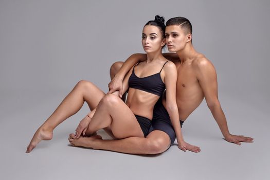 A pair of a graceful athletic ballet dancers in black suits are posing over a gray studio background. Good-looking male in black shorts is embracing a lovely woman in a black swimwear. Ballet and contemporary choreography concept. Art photo.