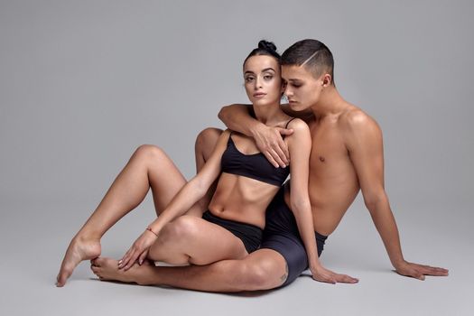 A pair of a young athletic ballet dancers in black suits are posing over a gray studio background. Attractive male in black shorts is embracing a beautiful woman in a black swimwear. Ballet and contemporary choreography concept. Art photo.