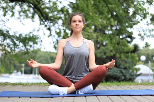 Cheerful slim female in tracksuit sitting on mat practicing yoga in lotus pose keeping calm in park