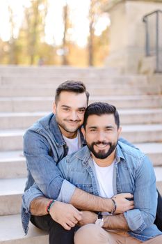 Caucasian handsome american young gays sitting on concrete stairs and hugging, wearing jeans shirts. Concept of same sex couple and lgbt.
