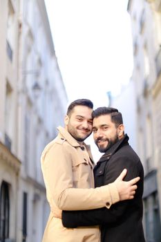 Two handsome caucasian hugging gays in city, buildings i background. Concept of same sex couple nad love.