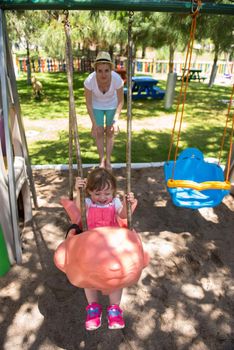 young mother and her little daughter smiling together while swinging at the playground in the park