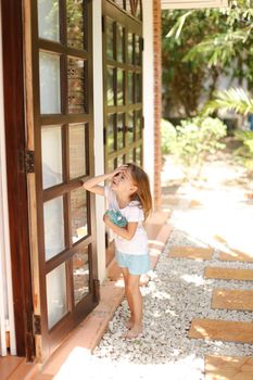 Little caucasian female kid standing near door with toy outside, palms in background. Concept of resting with kids in Phuket, exotic hotel.