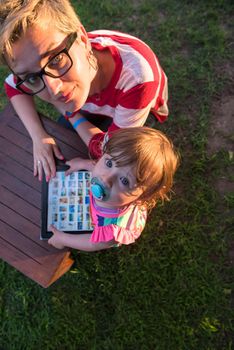 Happy mother and her little daughter enjoying free time using tablet computer while relaxing  on holiday home garden during sunny day