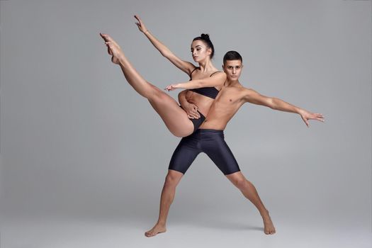 A couple of a graceful ballet dancers in black suits are posing over a gray studio background. Handsome man in black shorts is holding in his hands an attractive woman in a black swimwear. Ballet and contemporary choreography concept. Art photo.